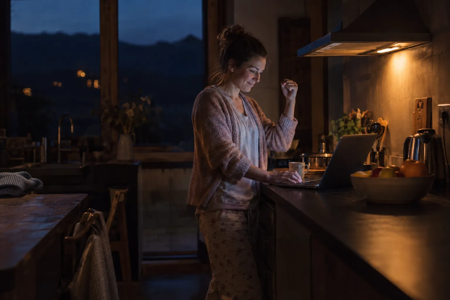 A woman in pajamas at a dark kitchen counter, typing on a laptop with one hand raised in a small victory fist.