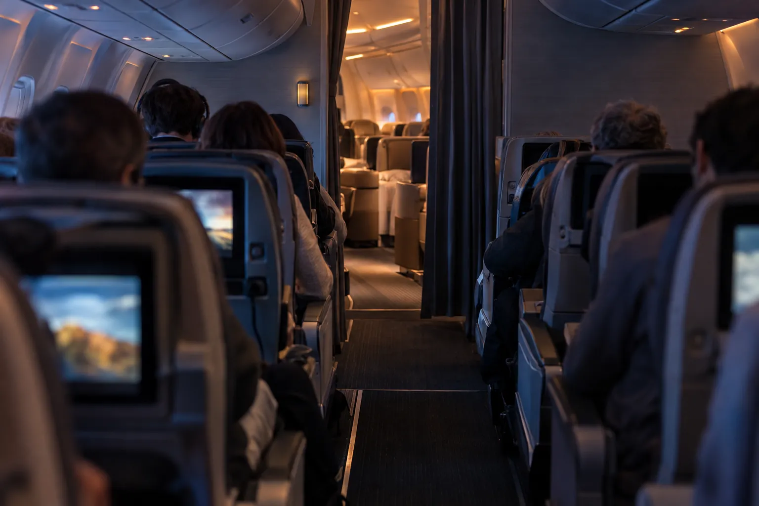 The aisle of a full economy cabin looking forward to a half-drawn curtain and a warmly lit business cabin beyond.