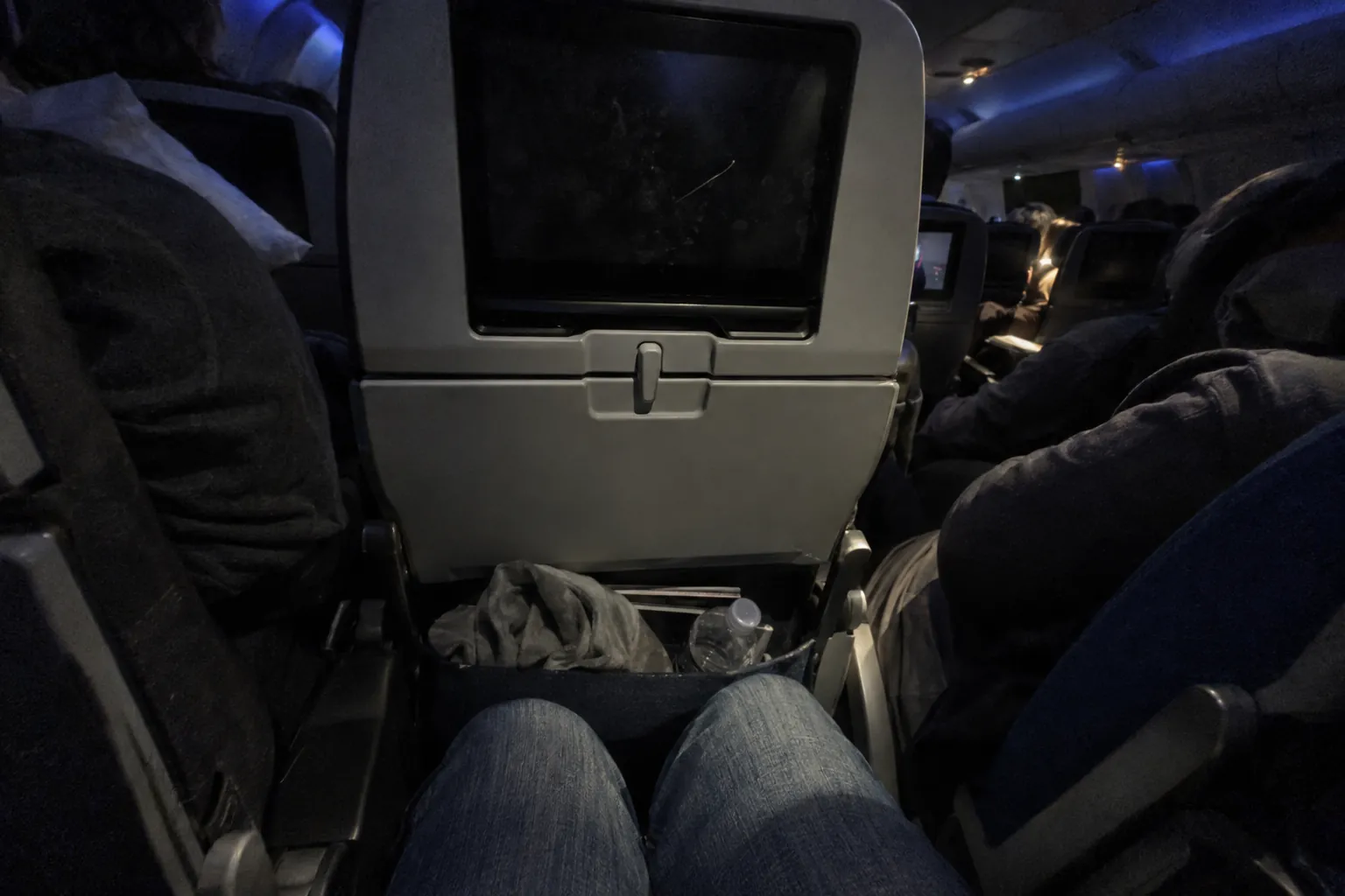 View from an economy middle seat at night: knees pressed against the seat in front, neighbors on both sides